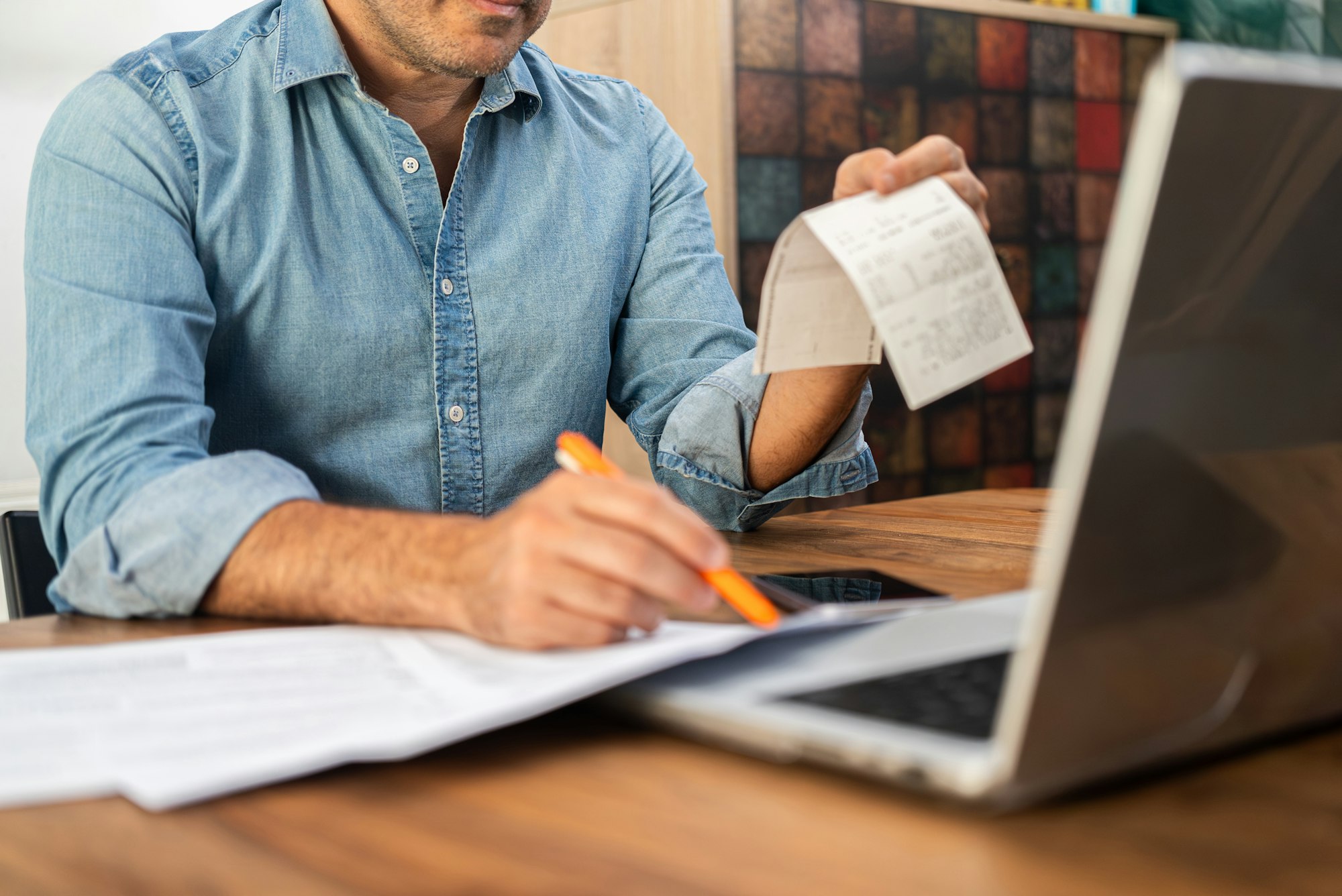 Middle-aged man calculating taxes at a wooden desk with a laptop and holding receipts.